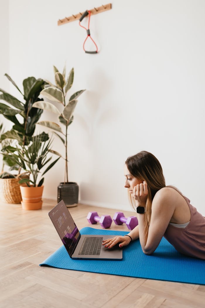 Serious young female in activewear lying on blue yoga mat on floor and browsing laptop